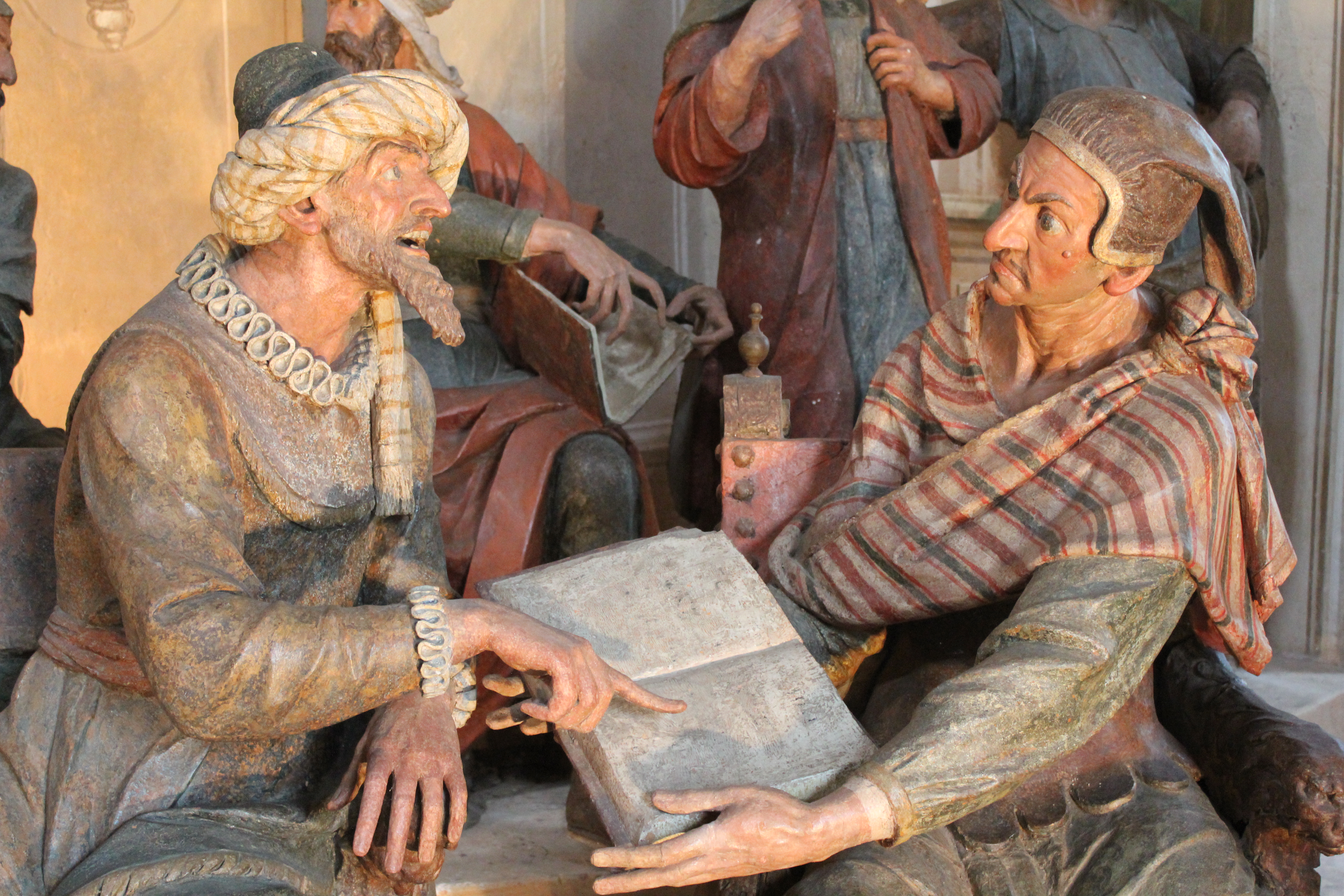 L'immagine raffigura una scultura dettagliata di due figure impegnate in una conversazione. Una figura, a sinistra, sembra essere un uomo con barba e capelli chiari fino alle spalle, vestito con abiti in stile medievale. Sta indicando un libro aperto tenuto dall'altra figura, che sembra essere una donna con un fazzoletto in testa e uno scialle a fantasia. Entrambe le figure appaiono attente e coinvolte, con espressioni e gesti vivaci, suggerendo uno scambio di conoscenze o una discussione.