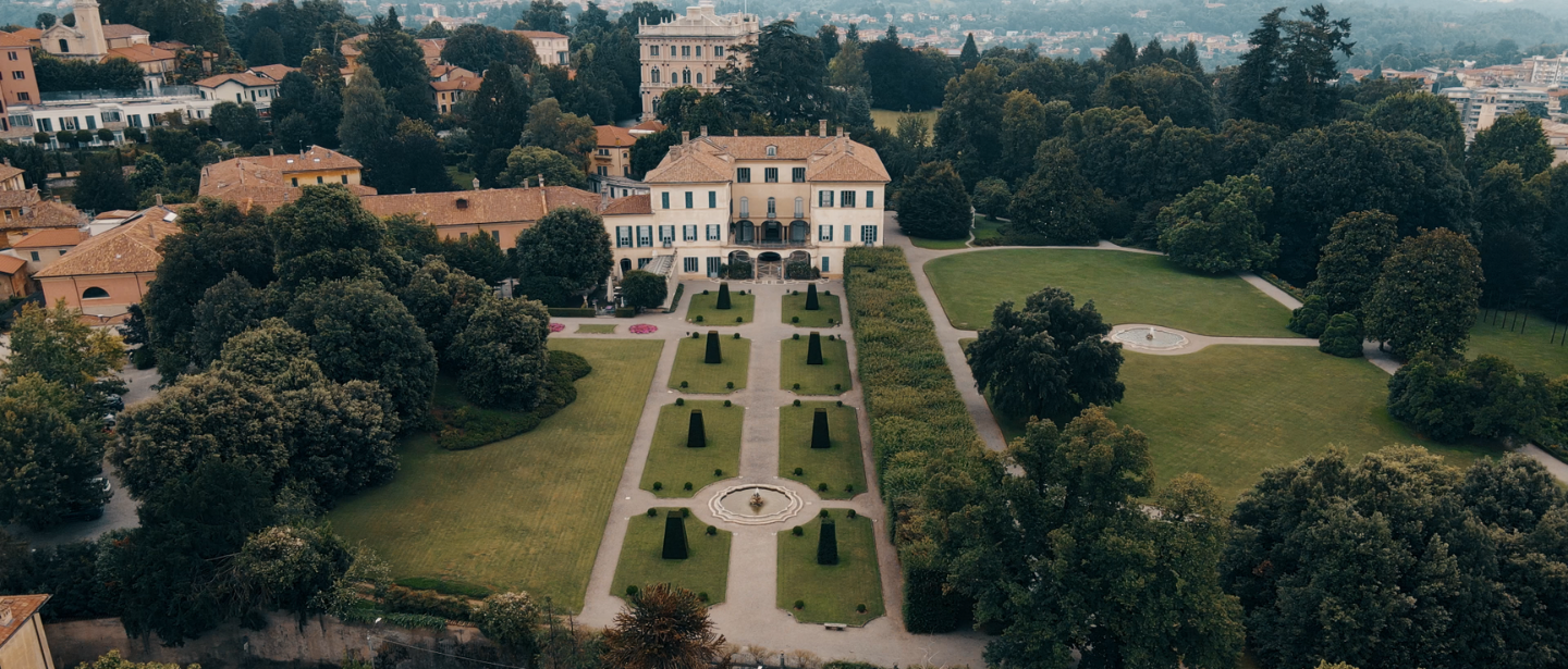 Fotografia aerea di Villa Panza a Varese, che mostra l'edificio principale con la sua elegante architettura neoclassica e il giardino geometrico curato. L'immagine include siepi, aiuole simmetriche, fontane e un ampio parco con alberi rigogliosi, circondato da edifici storici e il paesaggio urbano sullo sfondo.