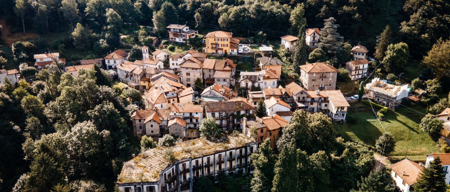 Immagine panoramica di un piccolo villaggio dipinto situato in mezzo alla natura, circondato da una fitta vegetazione. Gli edifici hanno tetti di tegole rosse e sono disposti in modo compatto lungo dolci pendii, creando un'atmosfera pittoresca e tranquilla.