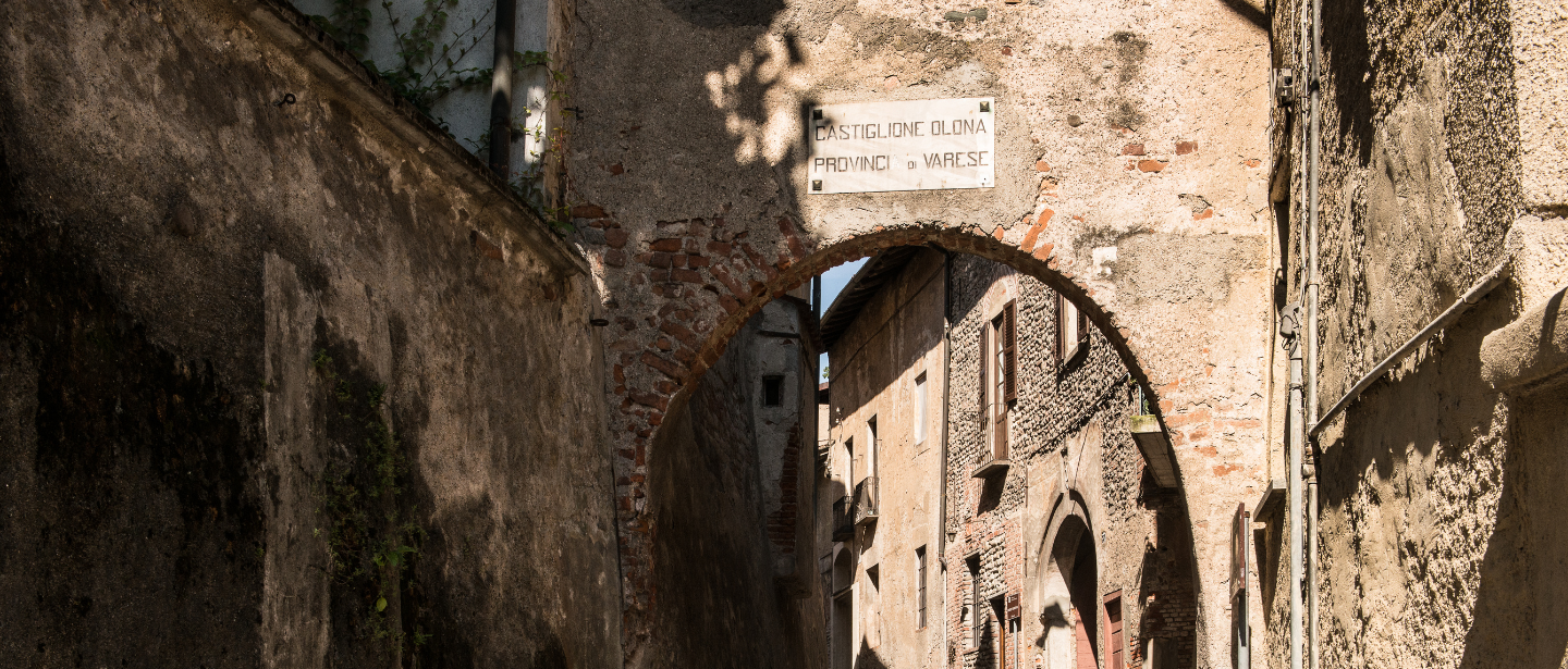 Veduta di un arco in pietra che segna l'ingresso al centro storico di Castiglione Olona, con una stretta via pavimentata e antichi edifici in pietra e mattoni.