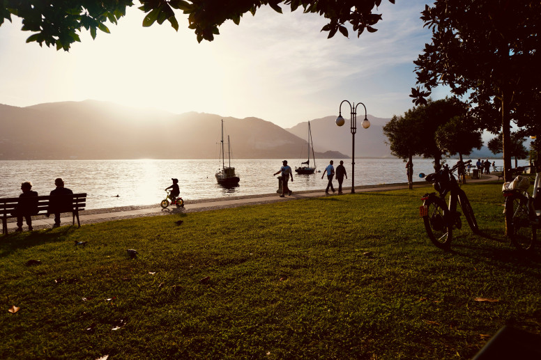 Lido erboso con lago e montagne e persone che camminano e fanno attività all'ora del tramonto.