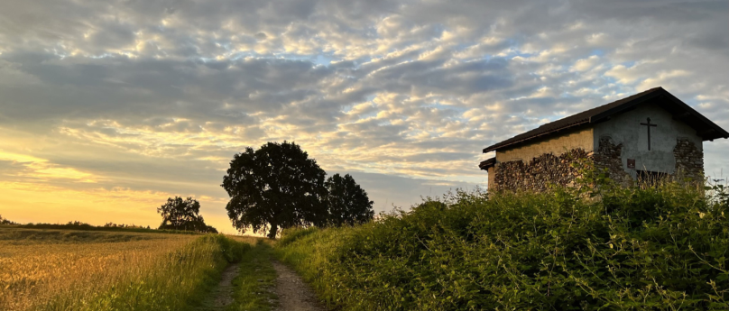 Un sentiero sterrato attraversa un paesaggio rurale al tramonto, circondato da campi dorati e vegetazione verde. A destra si trova una piccola cappella di pietra con una croce sulla facciata, mentre a sinistra si intravedono grandi alberi sotto un cielo nuvoloso illuminato dai caldi colori del tramonto. L'atmosfera è tranquilla e serena, con il contrasto tra la luce dorata e le ombre della sera che si allungano.