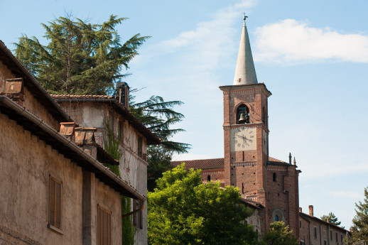 View of the brick bell tower of the Castiglione Olona church with the clock, surrounded by ancient houses and green trees under a clear sky.