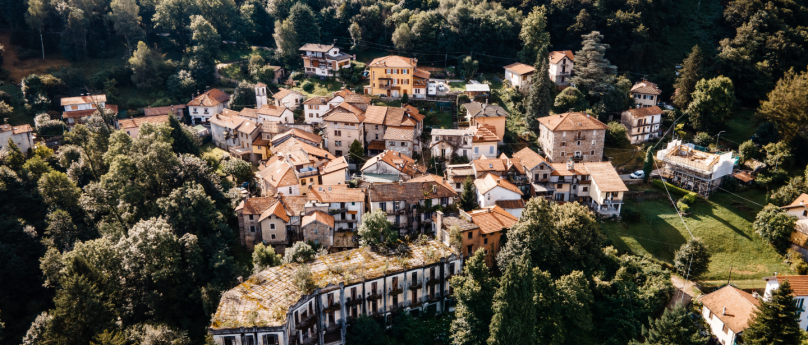 A panoramic image of a small painted village nestled in nature, surrounded by lush vegetation. The buildings have red-tiled roofs and are compactly arranged along gentle slopes, creating a picturesque and peaceful atmosphere.