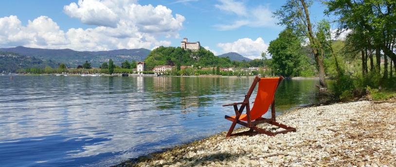 Lakeside promenade of Angera, with a small beach. By the lake, there is an empty sun lounger, and you can see the Rocca in the distance. 