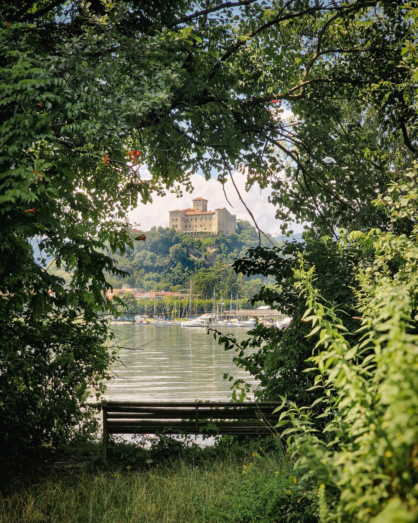 iew of the Rocca di Angera from the lakeshore. The fortress, at the center of the shot, is framed by green plants in the foreground. Between them, you can see the lake with sailboats gliding across it.
