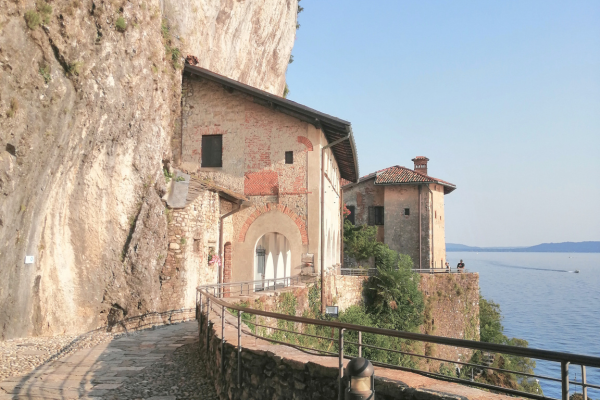 Stone structures built into the rock form a passage with arches. The path is bordered by a low wall and overlooks Lake Maggiore.